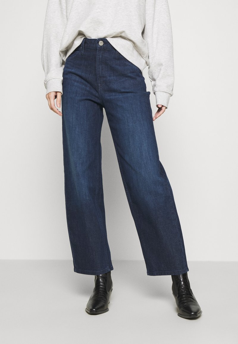 Osoba ve světlém šedém dlouhém rukávu, s tmavě modrými vysokými širokými džíny a černými kotníčkovými botami, stojící na jednobarevném pozadí.