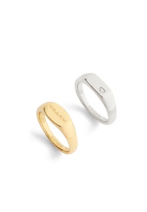 Due anelli signet di Coach, uno d'oro inciso con la scritta "COACH" e uno d'argento con un emblema a stella e "COACH" all'interno della fascia.