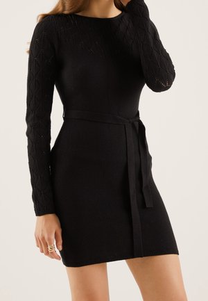 Model wears a fitted black knit dress with long patterned sleeves and a tied waist. She holds her hair back with one hand.