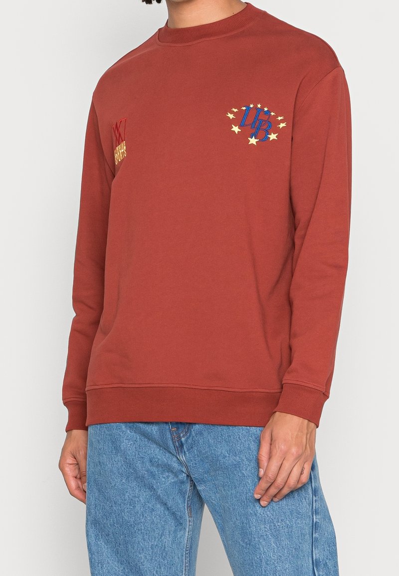 Homme portant un sweat-shirt couleur rouille avec un logo étoile bleu et jaune ainsi que du texte, associé à un jean en denim bleu clair, debout devant un fond blanc.