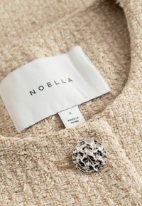 Tissu beige texturé avec un bouton décoratif argenté et une étiquette blanche indiquant la marque Noella et la taille small.
