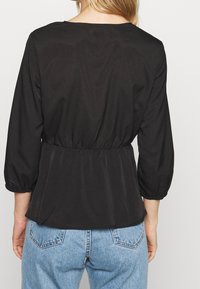 Top nero a maniche lunghe con scollatura rotonda e vita elasticizzata, caratterizzato da una texture liscia e maniche arricciate. Indossato con jeans blu.