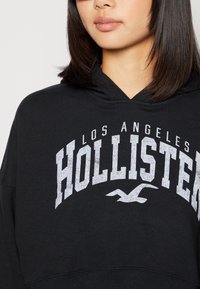 Felpa con cappuccio nera con una grande grafica bianca "HOLLISTER" e il testo "LOS ANGELES" sopra, orlo a coste e materiale morbido in misto cotone.