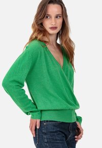 Pull vert en maille douce et texturée avec un col en V et des manches longues, présentant une coupe décontractée et un ourlet côtelé, associé à un jean foncé.