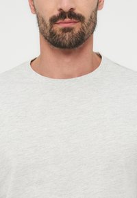 Άνδρας με σκούρα γενειάδα που φοράει ανοιχτό γκρι μπλουζάκι με στρογγυλή λαιμόκοψη, εμφανίζεται από τον λαιμό μέχρι το πάνω μέρος του στήθους, με φόντο απλό.