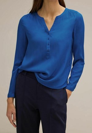 Frau trägt eine langärmelige blaue Bluse mit Knöpfen und dunkle Hose, steht mit einer Hand in der Tasche vor neutralem Hintergrund.