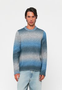 Pull en tricot avec un design dégradé dans des nuances de bleu et de gris, col rond, et poignets et ourlet côtelés, présenté avec un jean bleu clair.