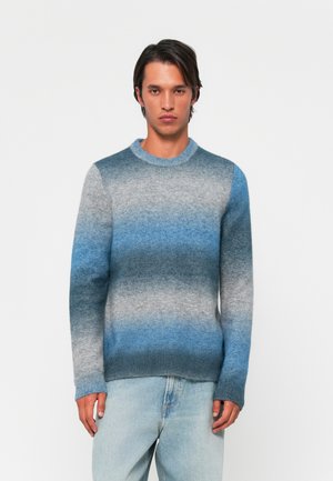 Strikket sweater med gradientdesign i nuancer af blå og grå, rund halsudskæring samt ribbede manchetter og søm, vist sammen med lyseblå jeans.