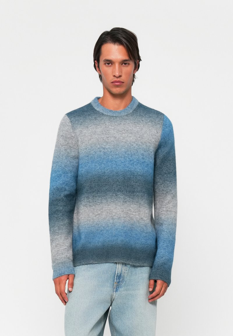 Pull en tricot avec un design dégradé dans des nuances de bleu et de gris, col rond, et poignets et ourlet côtelés, présenté avec un jean bleu clair.