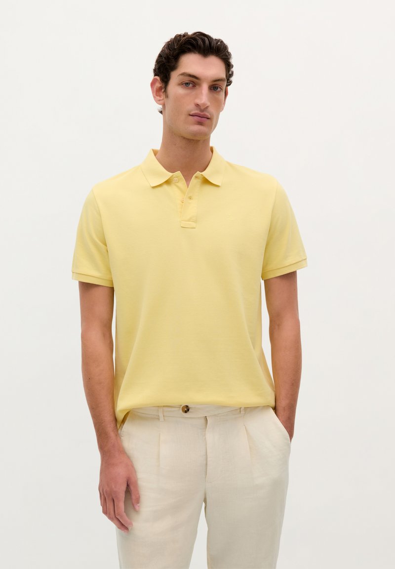 Gul polo skjorte med korte ærmer, let stof, to-knap lukning, parret med lysebeige bukser. Simpelt, rent design.