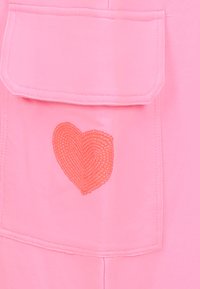 Roze stof met een voorkiep waar een geborduurd oranje hartontwerp op staat. Gladde textuur met gestikte details rond de zak.