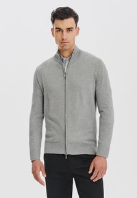 Szary sweter zapinany na zamek, wykonany z miękkiego materiału, z wysokim kołnierzem i ściągaczami przy mankietach. Noszony na jasnej koszuli, w połączeniu z ciemnymi spodniami.