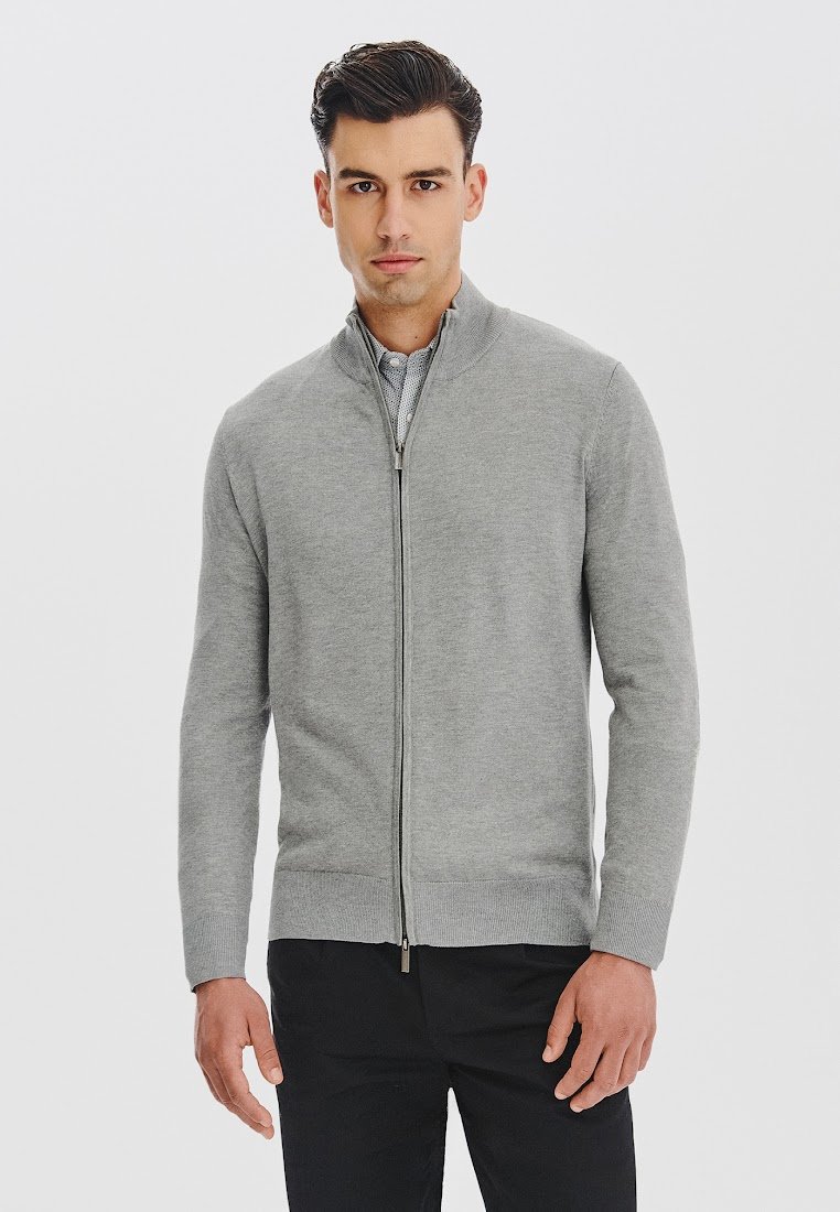 Szary sweter zapinany na zamek, wykonany z miękkiego materiału, z wysokim kołnierzem i ściągaczami przy mankietach. Noszony na jasnej koszuli, w połączeniu z ciemnymi spodniami.