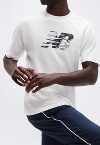 Μαύρο άτομο που φοράει λευκό t-shirt New Balance και ναυτικό παντελόνι με λευκή ρίγα, εμφανίζεται από τον λαιμό μέχρι το μέσο του μηρού, με τα χέρια σταυρωμένα μπροστά.