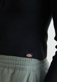 Svart ribbad tröja med en liten Dickies-logotyp ovanför midjebandet på lösa, ljusgrå byxor.
