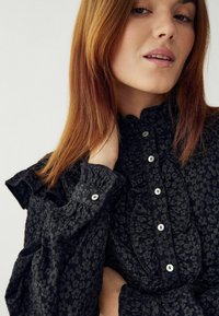 Czarna koszula w kwiaty z falbankami przy kołnierzu i rękawach, z małymi białymi guzikami i teksturowanym materiałem. Zbliżenie na wzór i detale.