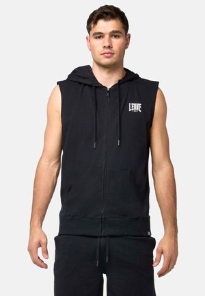 Jonge man staat, draagt een zwarte mouwloze zip-up hoodie met capuchon en een wit "LEONE 1947" logo op de borst.