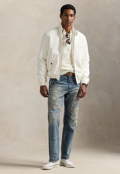 Vyras dėvi baltą bombonešį, kreminės spalvos pintinį megztinį, nusidėvėjusius mėlynus džinsus, baltus sportinius batelius, saulės akinius kabančius ant megztinio apykaklės.