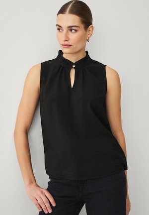 Kobieta z zaczesanymi do tyłu włosami, ubrana w czarny, bezrękawnik z falbanką przy dekolcie i zapięciem na guziki w kształcie kluczyka, pozująca na tle szarej ściany.