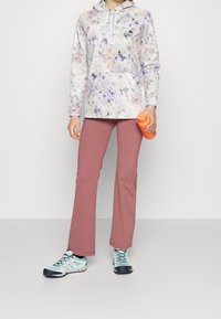 Pull à capuche légèrement imprimé dans des nuances de rose pastel et de violet, associé à un pantalon rose élastique et des chaussures de randonnée aqua, tenant une bouteille orange.