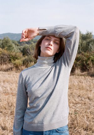 Une femme en pull gris se tient dans un champ sec et lève le bras au-dessus de son front pour se protéger de la lumière du soleil.