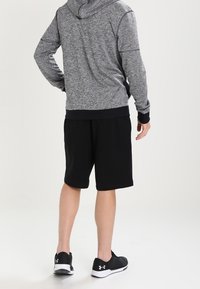 Mężczyzna w szarej bluzie z kapturem, czarnych szortach i czarnych butach sportowych marki Under Armour, stoi z plecami do aparatu.