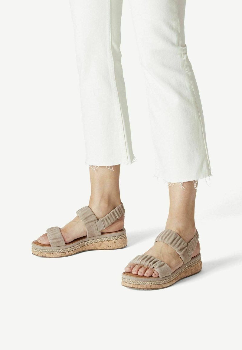 Pieds chaussés de sandales compensées beiges à brides froncées, associés à un pantalon blanc court à ourlet effiloché.