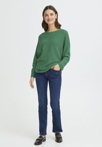 Pull vert en maille avec un large col, coupe décontractée et tissu texturé, associé à un jean bleu et des mocassins noirs ornés d'accents dorés.