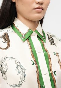 Γυναίκα που φοράει κρεμ πουκάμισο με πράσινο περίγραμμα και τυπωμένα ζώα, όπως ψάρι, σαύρα και καμηλοπάρδαλη, ορατά κοντά στον γιακά.