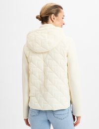 Helle, gesteppte Jacke mit Kapuze, die über Rautenmuster, eine weiche Textur und lange, taillierte Ärmel verfügt, kombiniert mit blauen Jeans.