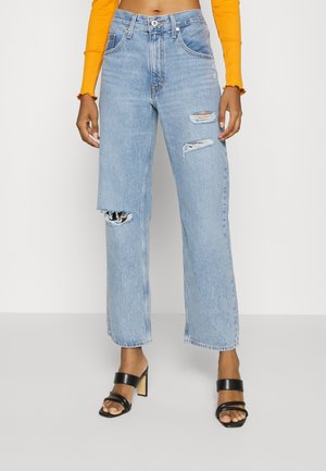Jean en denim bleu clair avec une coupe droite, des détails usés et un ourlet large ; associé à des sandales à talons strappy noires.