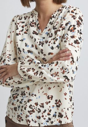 Kvinde iført en cremefarvet bluse med brune, sorte og blå abstrakte pletter, korslagte arme, synlig fra mellemtorsoen.