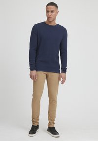 Mornarsko pleteno dolgimi rokavi pulover, svetlo rjave slim-fit hlače in črne športne copate. Pulover ima okroglo izrez in teksturirano tkanino.