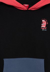Hoodie noir avec une capuche intérieure rouge. Présente un graphique d'une bouche ouverte sur la poitrine. Poche bleue à l'avant. Fabriqué en tissu en mélange de coton.
