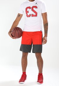 Röda och svarta atletiska shorts med meshtäckning, tillsammans med en vit t-shirt som har en stor röd "23" grafisk design, hållande en basketboll.