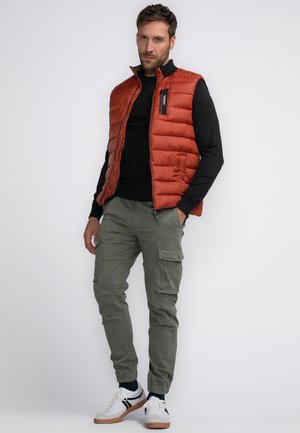 Orange padded vest over a black long-sleeve shirt, olive cargo pants, and white sneakers with black accents; standing against a white background.