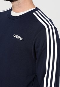 Sudadera azul marino con diseño de tres rayas blancas en los hombros y logo de "adidas" en blanco en el pecho. Hecha de material suave y con cuello redondo.