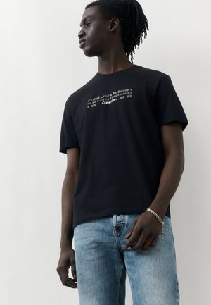 Jeune homme à la peau foncée et aux dreadlocks, portant un t-shirt noir Calvin Klein et un jean bleu clair, debout devant un fond blanc uni.