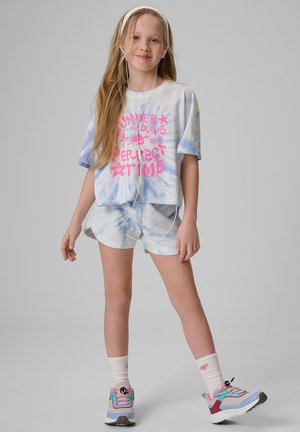 Uśmiechnięta dziewczyna w koszulce i szortach tie-dye z napisem „Summer Days Perfect Time”, w słuchawkach, sportowych butach i białych skarpetkach, stoi.