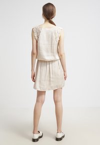 Robe beige à coupe ample, avec des accents en dentelle sur les épaules, texture lisse et longueur courte, associée à des chaussures blanches.