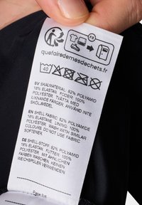 Étiquette d'entretien en tissu blanc fixée à un vêtement noir, présentant les instructions d'entretien et la composition du matériau en plusieurs langues.