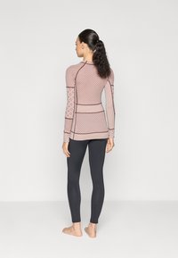 Haut ajusté à manches longues en rose clair avec des motifs géométriques et des coutures contrastées, associé à des leggings noirs. Tissu lisse et extensible, pieds nus.