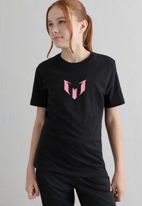 Mladý člověk s červenými vlasy svázanými vzadu, oblečený v černém tričku s růžovým abstraktním logem, stojící před jednobarevnou bíle texturovanou zdí.