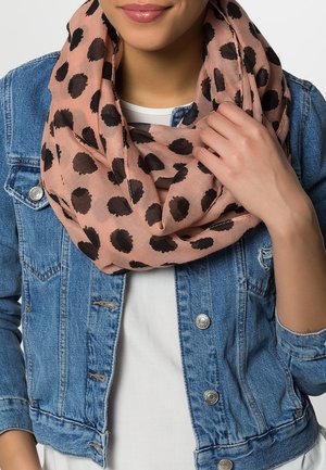 Femme portant une écharpe rose clair à pois noirs sur une chemise blanche et une veste en jean bleue, tenant l'écharpe près de l'épaule.