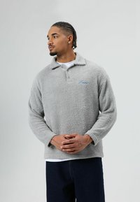 Fleece-pullover in lichtgrijs met een kraag, lange mouwen en zakken; beschikt over een blauw geborduurd logo en een gestructureerde stof.