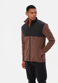 Giacca in pile marrone con una parte superiore nera, chiusura con zip completa e tasca con zip. Presenta un colletto alto e texture a contrasto.