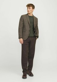 Blazer de tweed marrón con dos botones, suéter de punto verde debajo, pantalones oscuros marrones y zapatos negros de cordones. Fondo neutro.