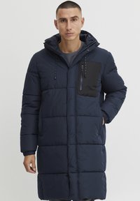Abrigo acolchado azul marino con un bolsillo en el pecho con cremallera, capucha y puños elásticos. Presenta un diseño acolchado y un corte de longitud media para mayor calidez.