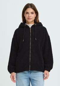 Veste en polaire noire avec fermeture éclair à l'avant, capuche et cordons de serrage. Elle a un extérieur texturé et duveteux et des manches amples.