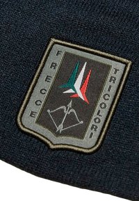 Cappello di maglia nero con un patch scudo ricamato. Il patch presenta un emblema stilizzato con elementi verdi, bianchi e rossi e un design a fiocco.